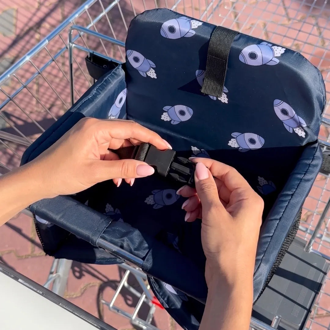 Coco Comfort Seat in navy blue astronaut pattern, hands buckling safety straps inside a shopping cart