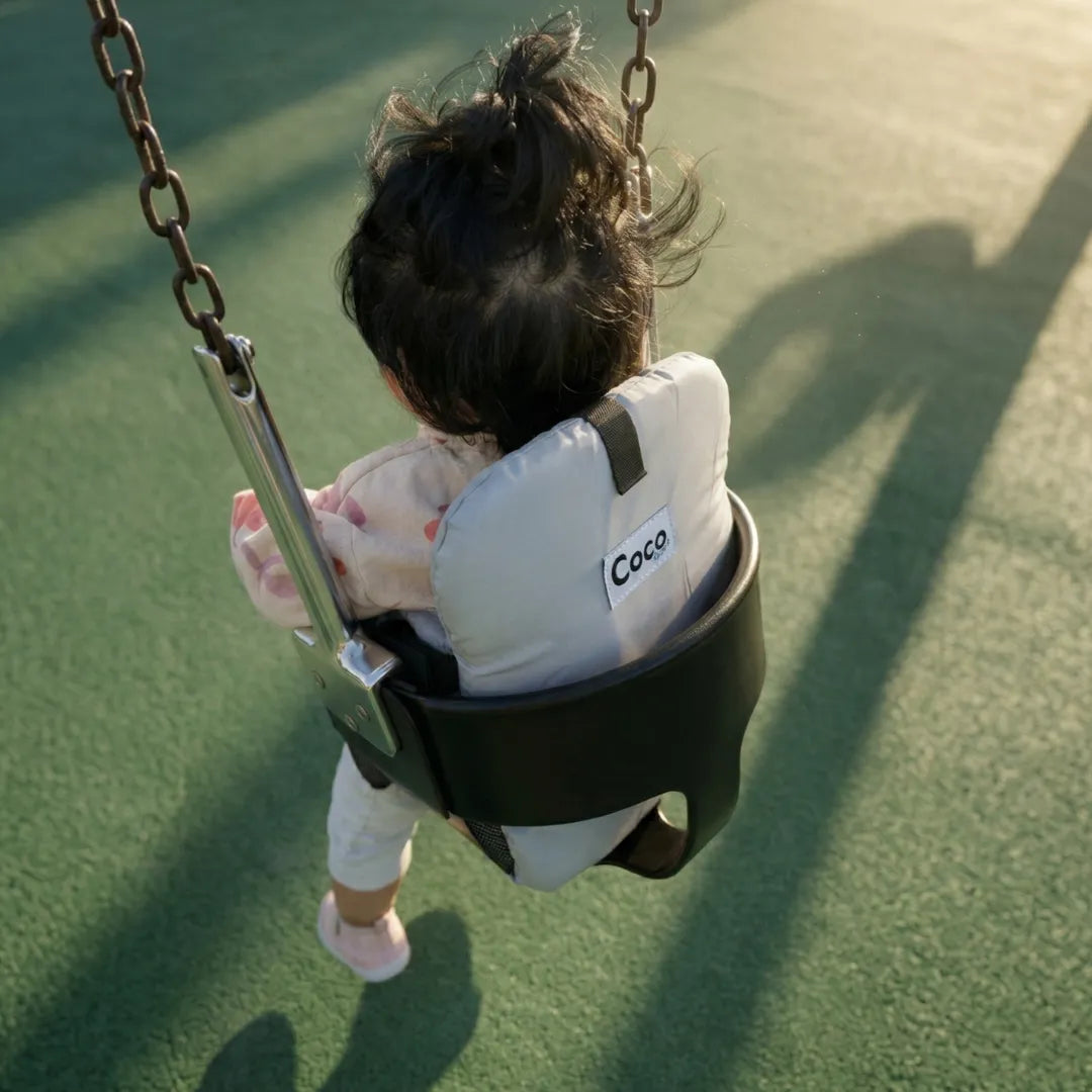 Baby in Cocoseat insert on playground swing, rear view showing padded back support and brand label